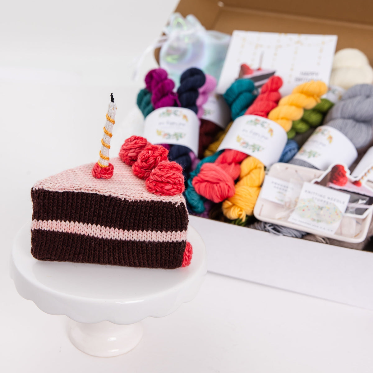 a hand knit slice of birthday cake sits on a white pedestal in front of a box full of birthday yarn bundles perfect for hand dyed yarn lovers. 