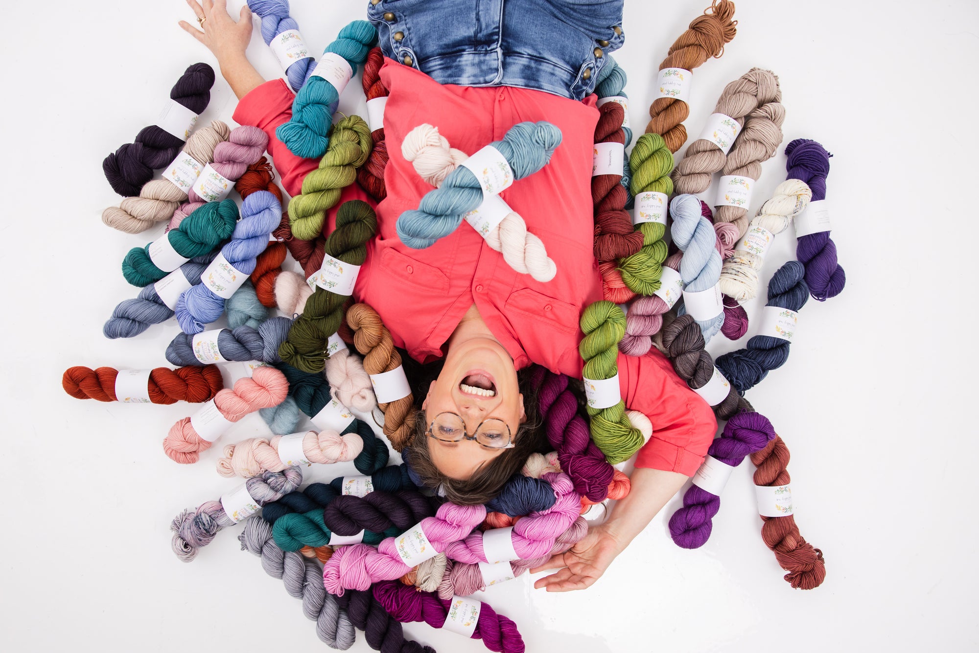 A woman is laying on a white background surrounded by skeins of yarn dyed in many different colors. She is laughing and wearing a bright coral shirt. She has brown hair and glasses.