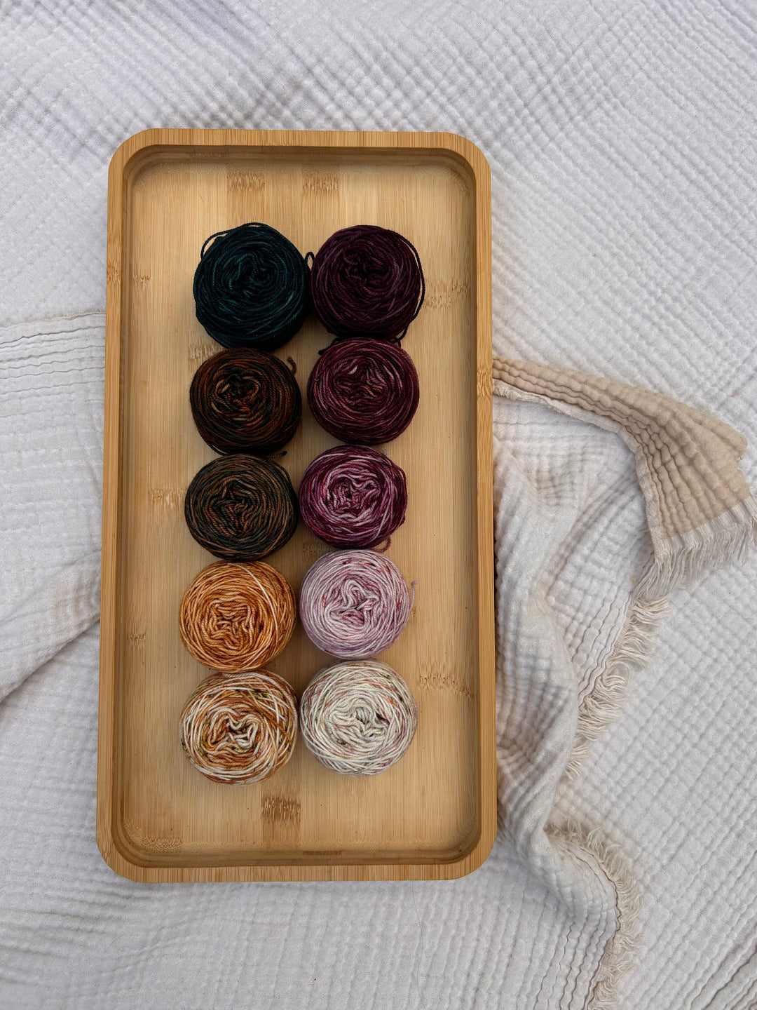 Ten cakes of yarn arranged in two rows on a rectangular bamboo tray, set against a textured white blanket with a cream fringed throw. The yarn colors include various shades of purple, brown, beige, and dark teal.