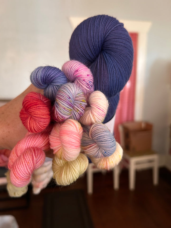 A hand holding a bundle of multiple variegated yarn skeins in different colors, including navy blue, purple, pink, red, and cream, with a blurred indoor room in the background.